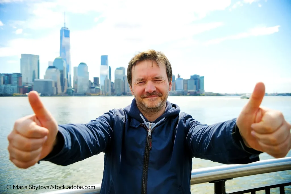Ein Hoschschul-Mitarbeiter vor einer amerikanischen Stadtsilhouette richtet beide Daumen nach oben