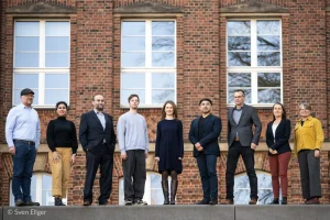 Das Team von Leonardo-Sachsen steht auf einer Mauer vor einer Backsteinfassade